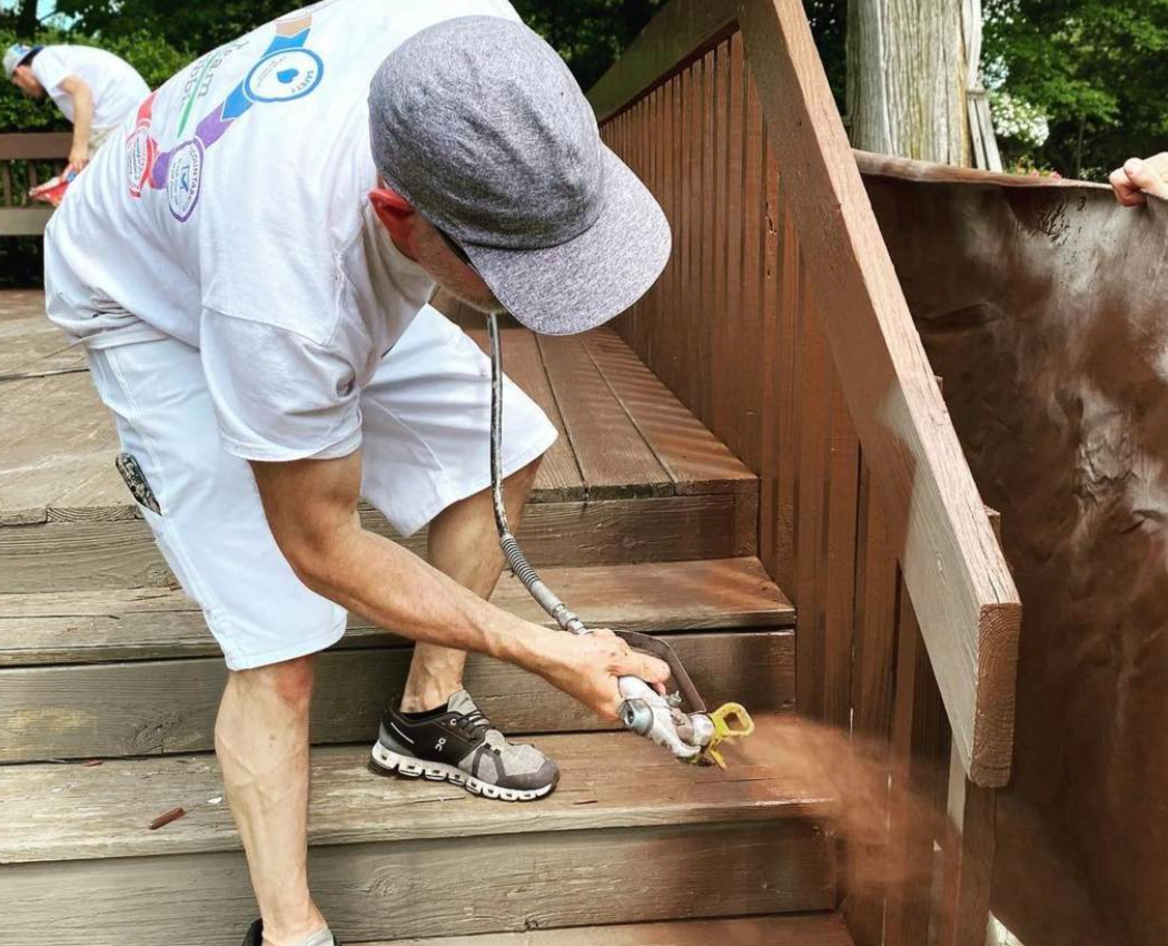 A painter sprays paint onto a bare wood deck