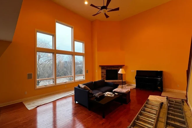 A spacious living room in Southeast Michigan—featuring orange walls, expansive windows, a sleek black sofa set, a black piano, fireplace, and a wooden ladder resting on gleaming hardwood floors—showcases the craftsmanship of Tribble Painting. Homes in communities like Ann Arbor, Ypsilanti, and Novi benefit from the expert touch that brings vibrant color and quality finishes to each room. by Tribble Painting