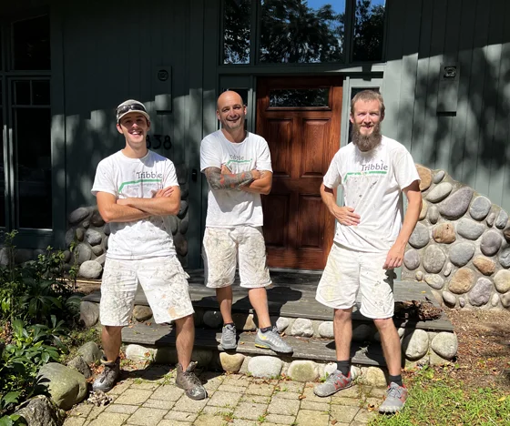 Three men in white painter uniforms stand smiling in front of a wooden door and stone wall exterior, proudly representing Tribble Painting and showcasing their residential painting expertise on a sunny day in Southeast Michigan communities such as Ann Arbor, Ypsilanti, and Novi. by Tribble Painting