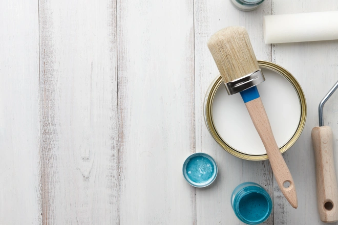 Paint brush, sponge roller, paints, waxes and other painting or decorating supplies on white wooden planks, top view