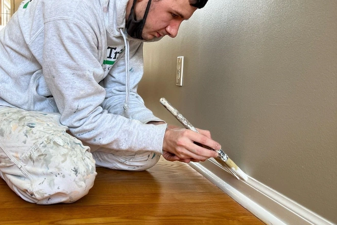 Tribble Painting Company employee carefully applying paint to a baseboard in a hallway with wood floors
